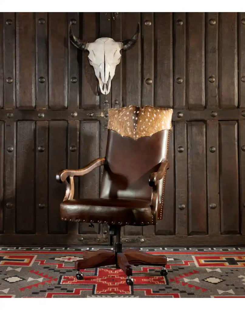 The Bowie Axis Dark Brown Desk Chair placed in a rustic interior, featuring supple pull-up leather, a hand-stitched Axis Deer Hide yoke, and a Southwestern-style rug beneath.