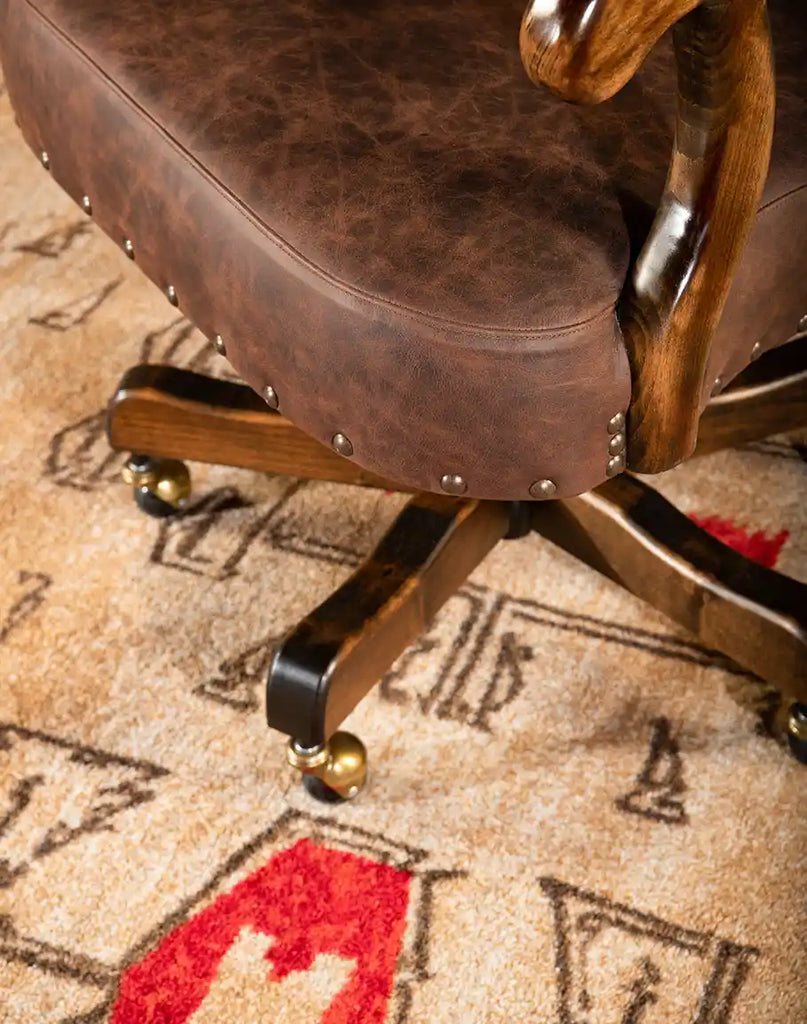 A close-up view of the Bowie Axis Taupe Desk Chair's base, featuring distressed taupe leather upholstery, decorative nailhead trim, and sturdy wooden legs with brass casters on a patterned rug.