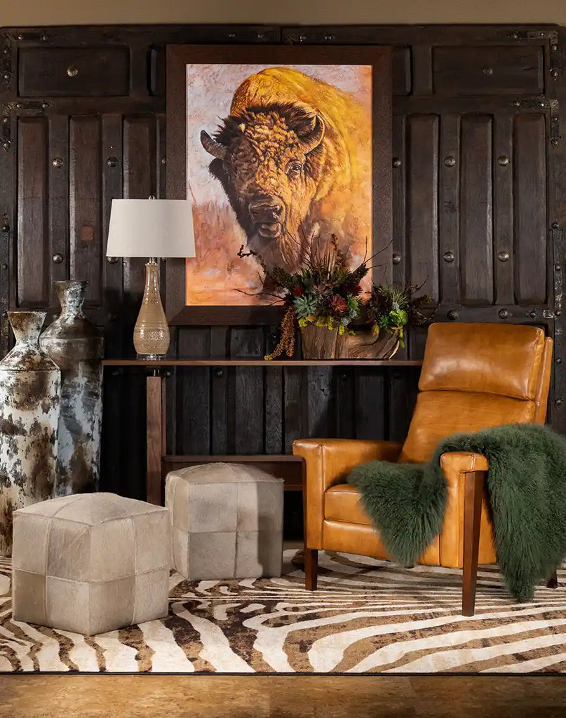 A cozy Western-inspired living space featuring a caramel leather recliner with a green fur throw, set against a dark wooden panel wall. A large framed bison painting, decorative vases, and a zebra-patterned rug complete the rustic yet elegant ambiance.