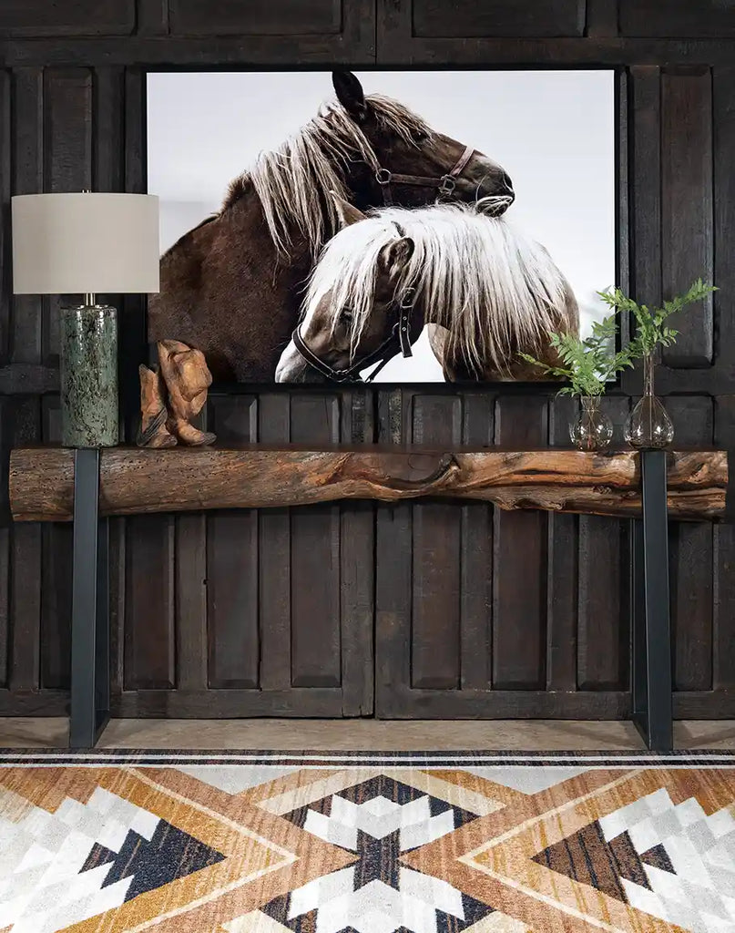 "The Tenderhearted" framed art featuring two horses in a tender embrace, displayed in a rustic interior with dark wood paneling, a natural wood console table, and decorative elements including a lamp and greenery.