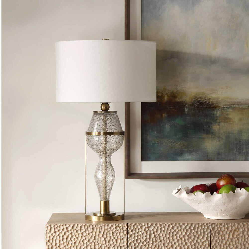 Venus Hourglass Table Lamp featuring a seeded glass base with gray flecks, antiqued brass accents, and a white linen drum shade.